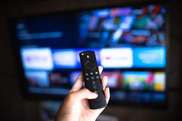 Hand holding an Amazon Fire TV remote in front of a television displaying streaming apps.