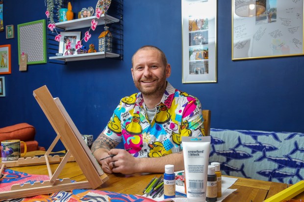 A man in a colorful shirt sits at a table, painting at an easel.