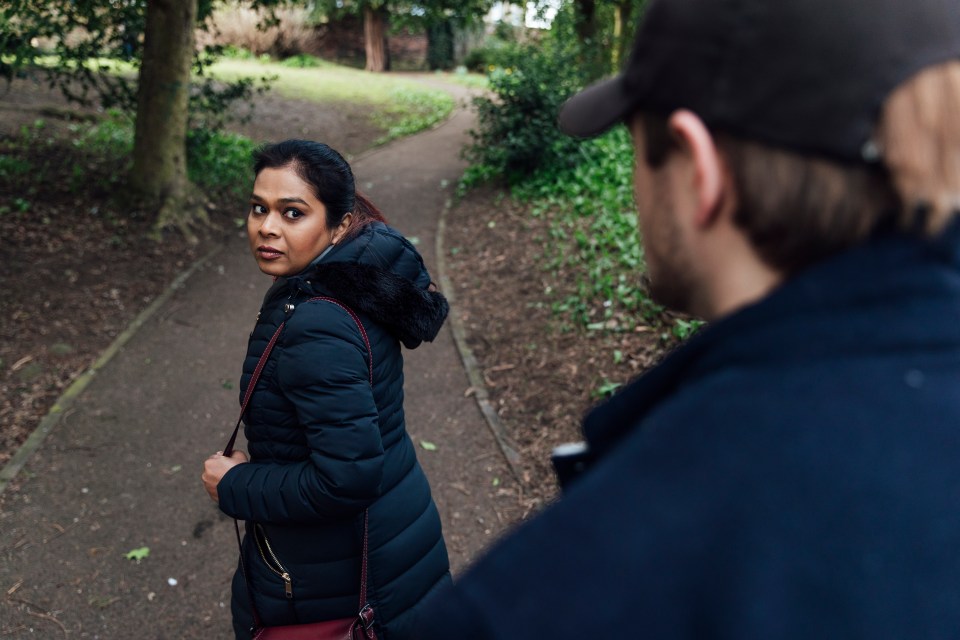 A terrified woman walking in a park looks over her shoulder at a man following her.