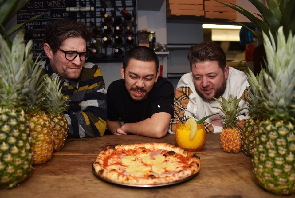 Three men looking at a Hawaiian pizza with pineapples.