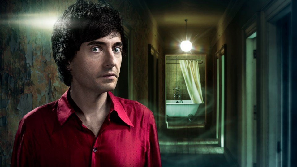 Danny Robins in a red shirt, standing in a dimly lit hallway.