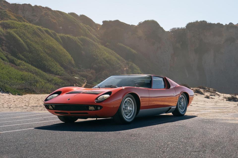 Red Lamborghini Miura P400 coupe driving on a road near a beach.