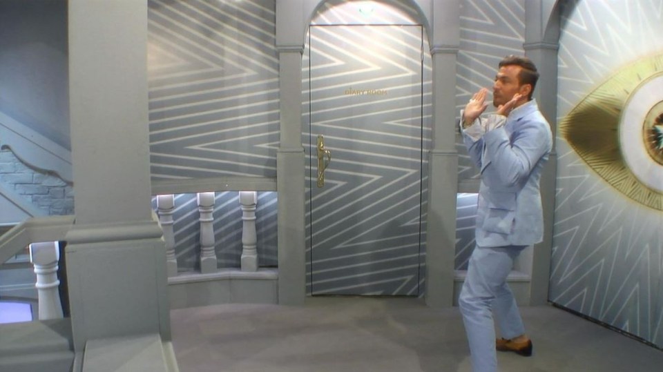 Paul Danan in a light blue suit standing before a diary room door.