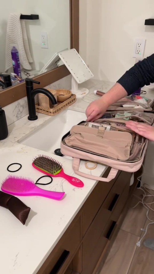 A person packing a travel case with toiletries and hair accessories on a bathroom counter.