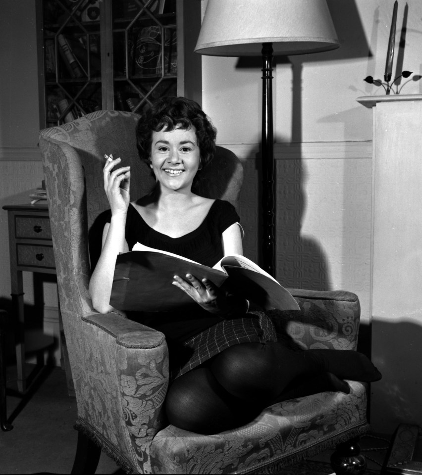 Black and white photo of Joan Plowright at home, holding a book and smoking a cigarette.