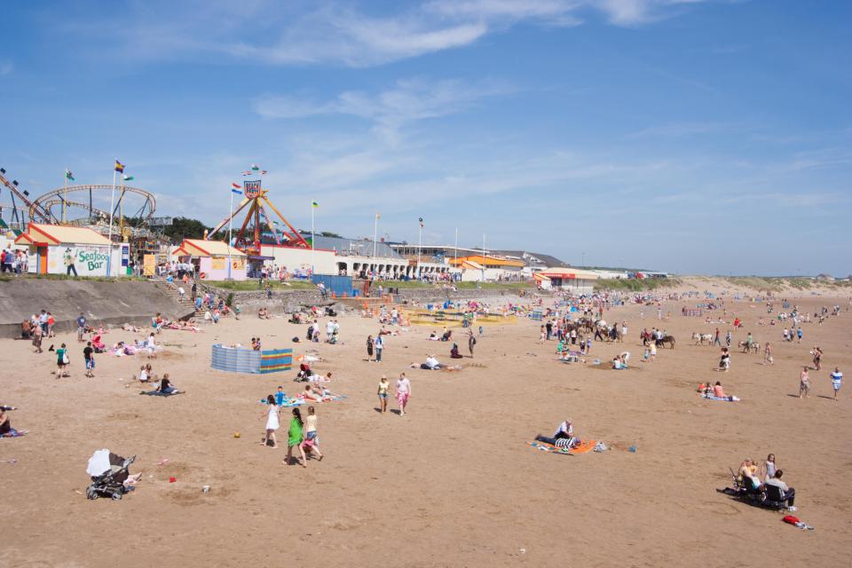 Crowded sandy beach with amusement park rides in the background.