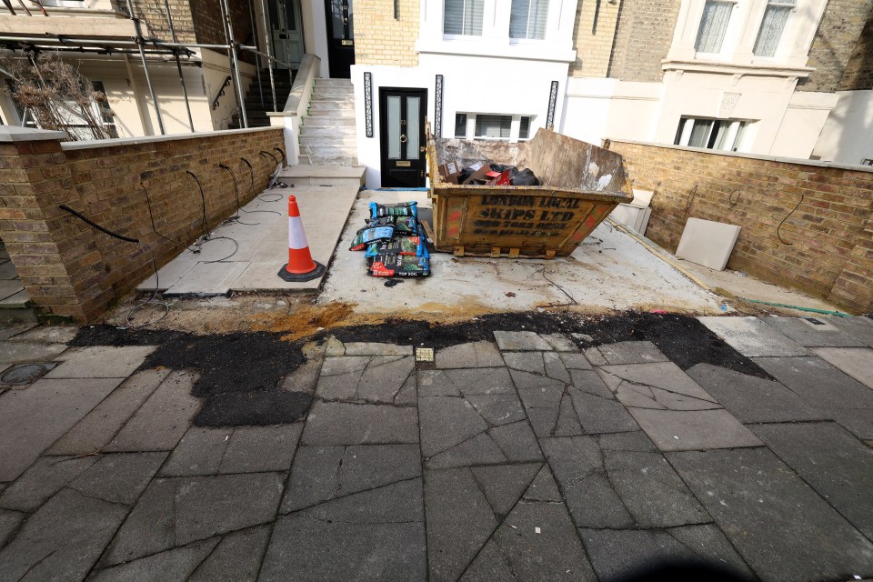 Damaged pavement outside a house under construction, with a skip and bags of soil.