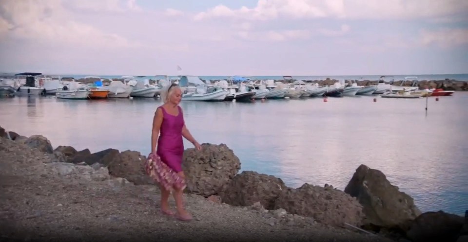 Woman in pink dress walking by the sea with boats in the background.