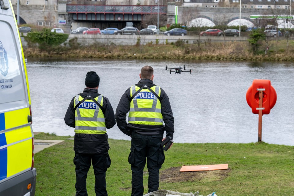 Police officers monitoring a drone over a river during a recovery operation.