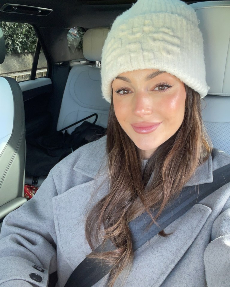 Michelle Keegan in a car, wearing a grey coat and white beanie.