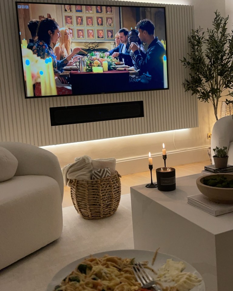 A television screen showing a group of people at a dinner table, with a cozy living room setting including a basket of blankets and a plate of food in the foreground.