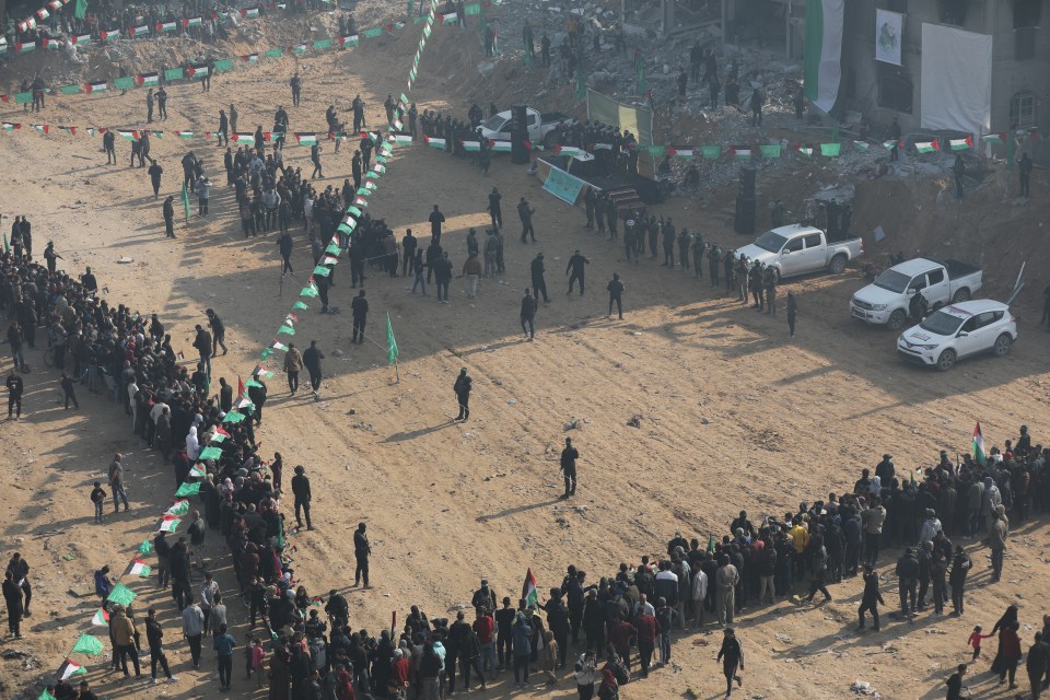 Aerial view of a large group of people gathered in a sandy area, many holding Palestinian flags.  Vehicles are present.