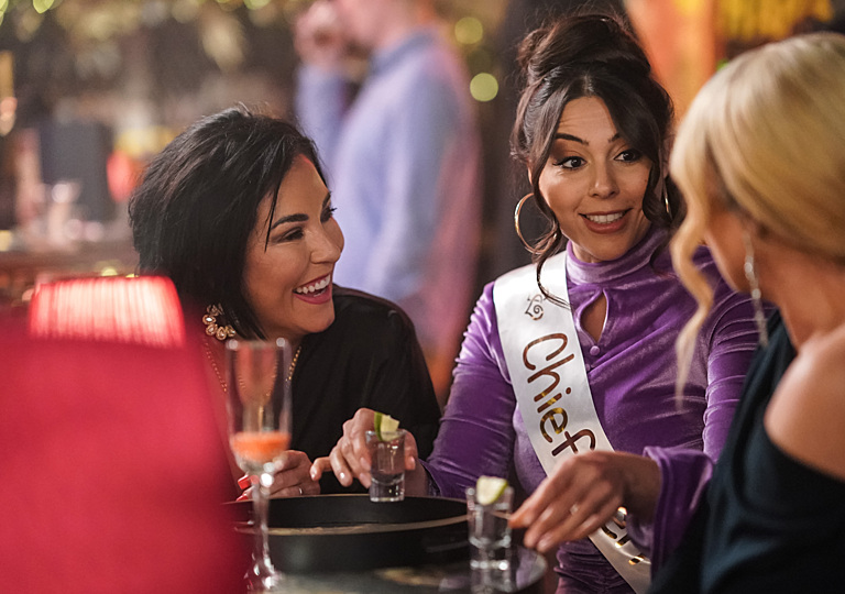 Three women at a bar; one is wearing a "Chief" sash.