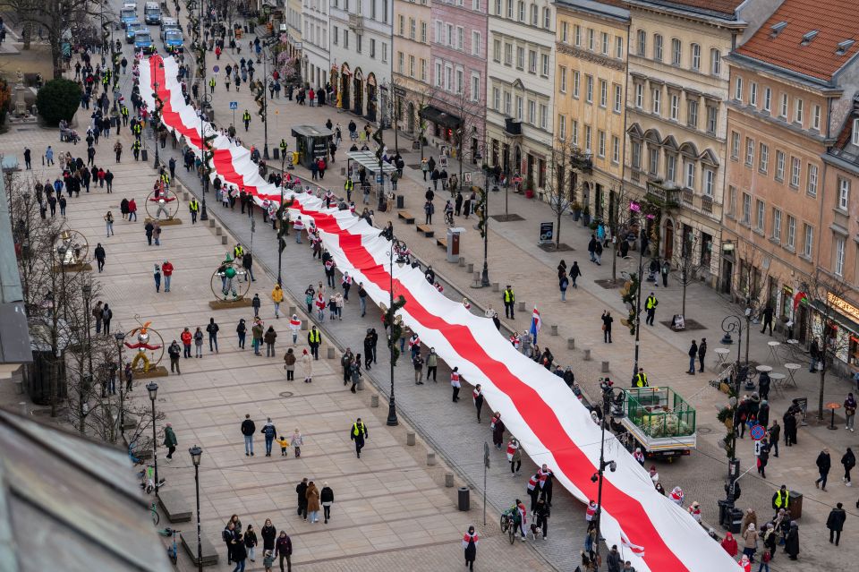 Protesters carrying a large historical Belarusian flag in a city street.