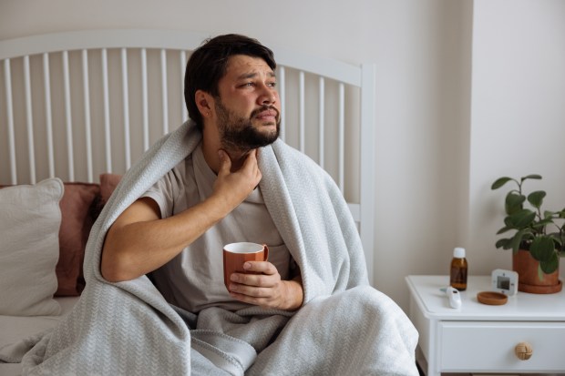 Man sitting in bed, holding a mug and touching his throat.