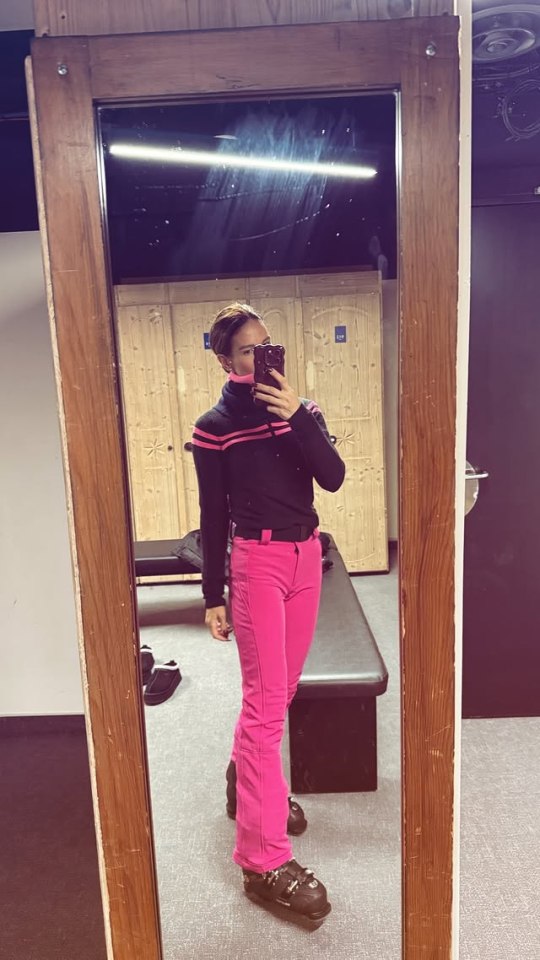 Woman in pink ski pants and black sweater taking a selfie in a mirror.