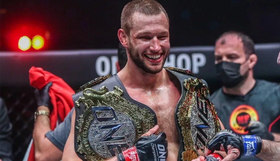 Reinier de Ridder smiling and holding two championship belts.