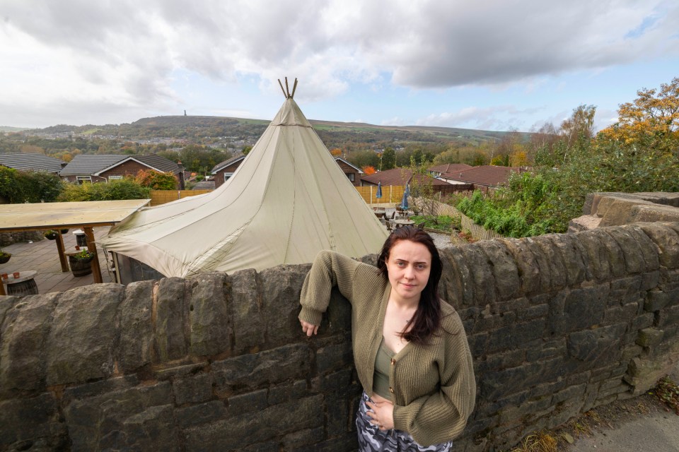 A woman leans on a stone wall in front of a teepee in a pub garden.