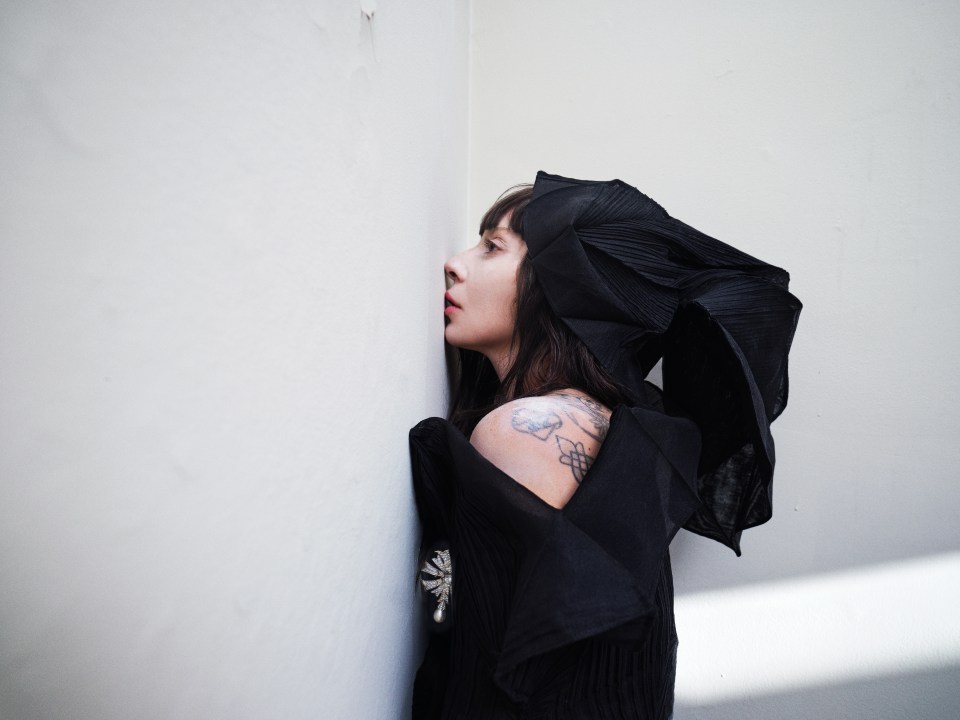 Lady Gaga, wearing a black head covering and top, leans against a white wall.