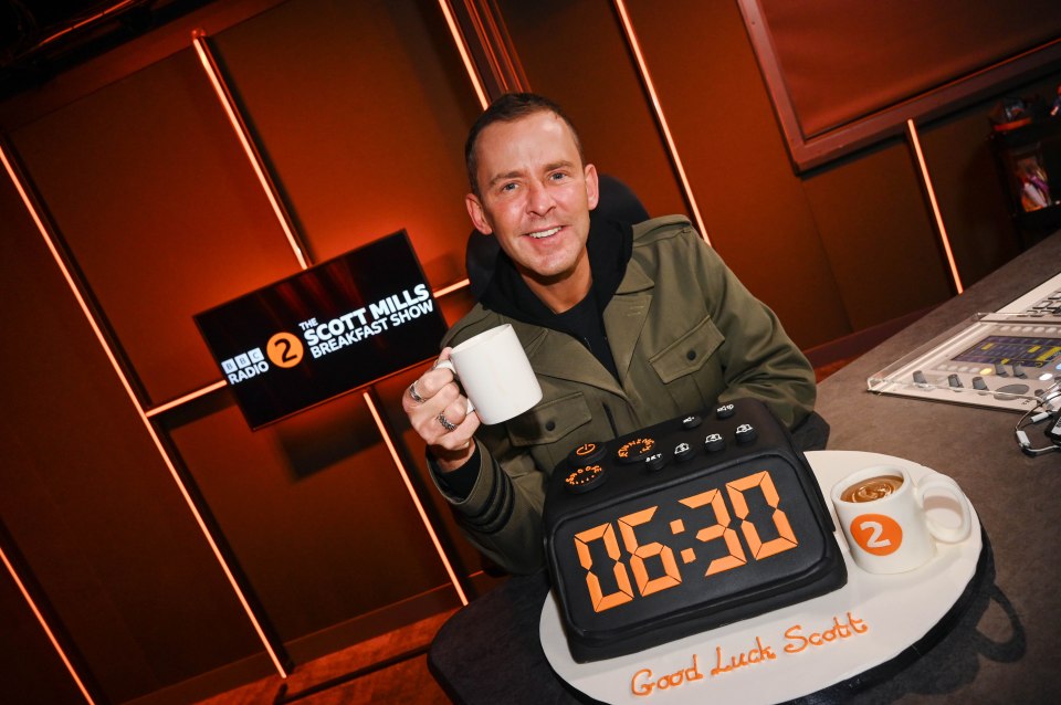 Scott Mills holding a mug, sitting in front of a clock cake for his first BBC Radio 2 Breakfast Show.