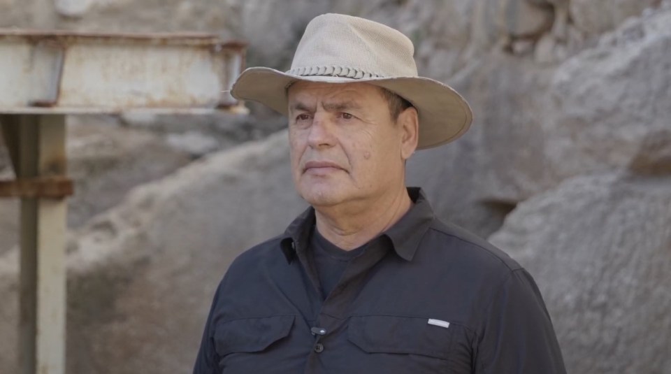 A man in a hat stands near a rocky outcrop.