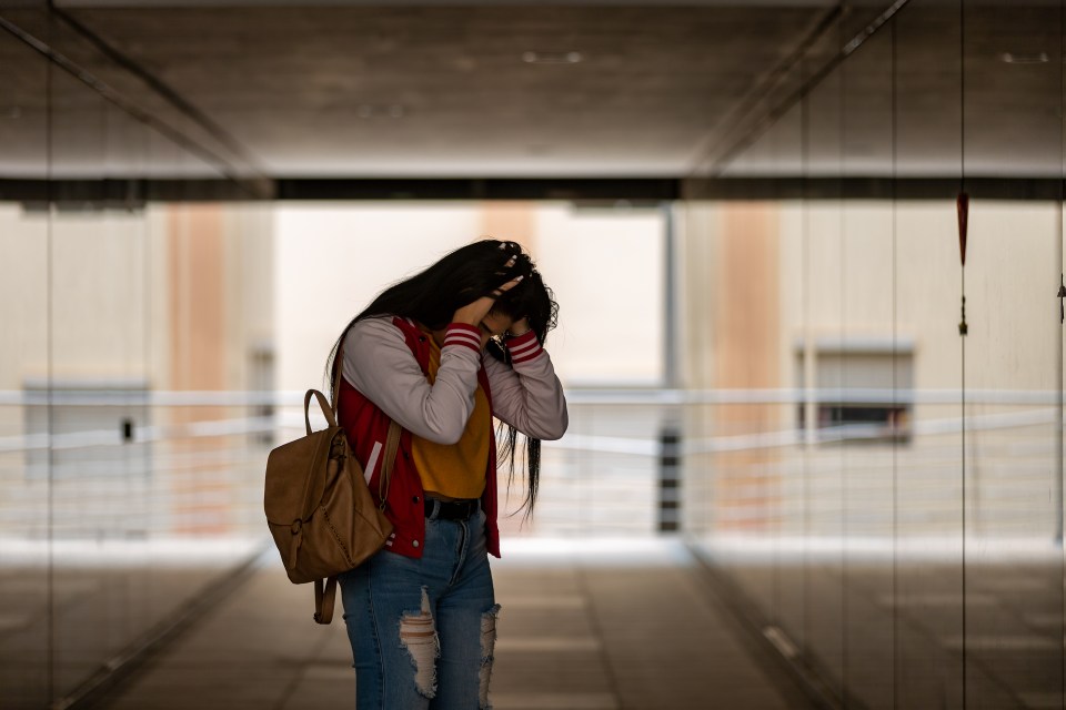 Teenage girl with backpack looking down, hands on head, appearing distressed.