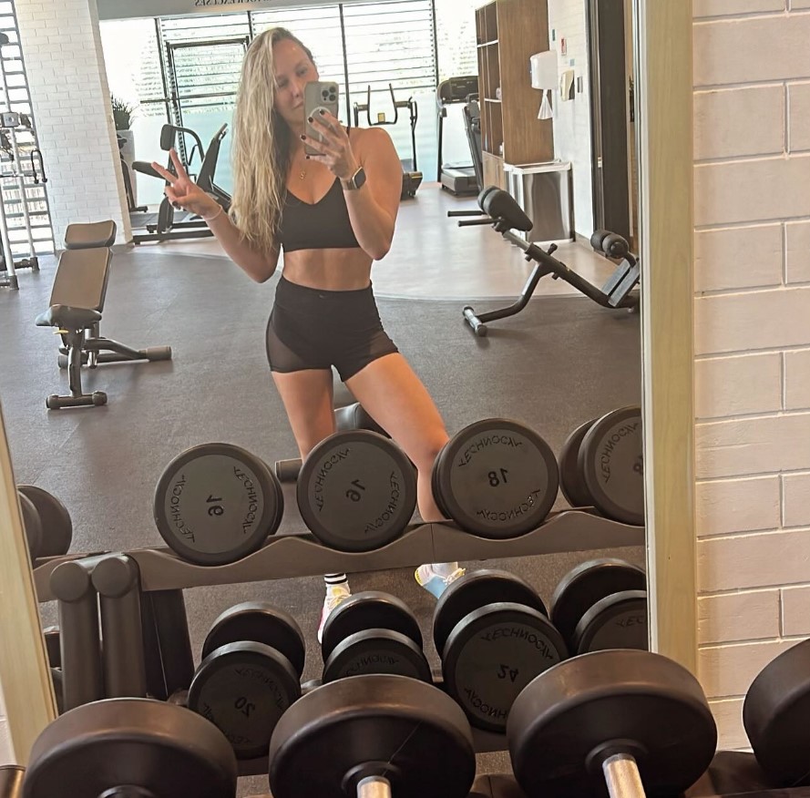 Woman in gym selfie, showing peace sign.