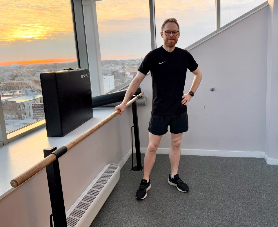 Man in workout clothes standing by a barre in a gym with a city view.