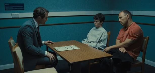 Scene from a crime drama showing three people sitting around a table in an interrogation room.