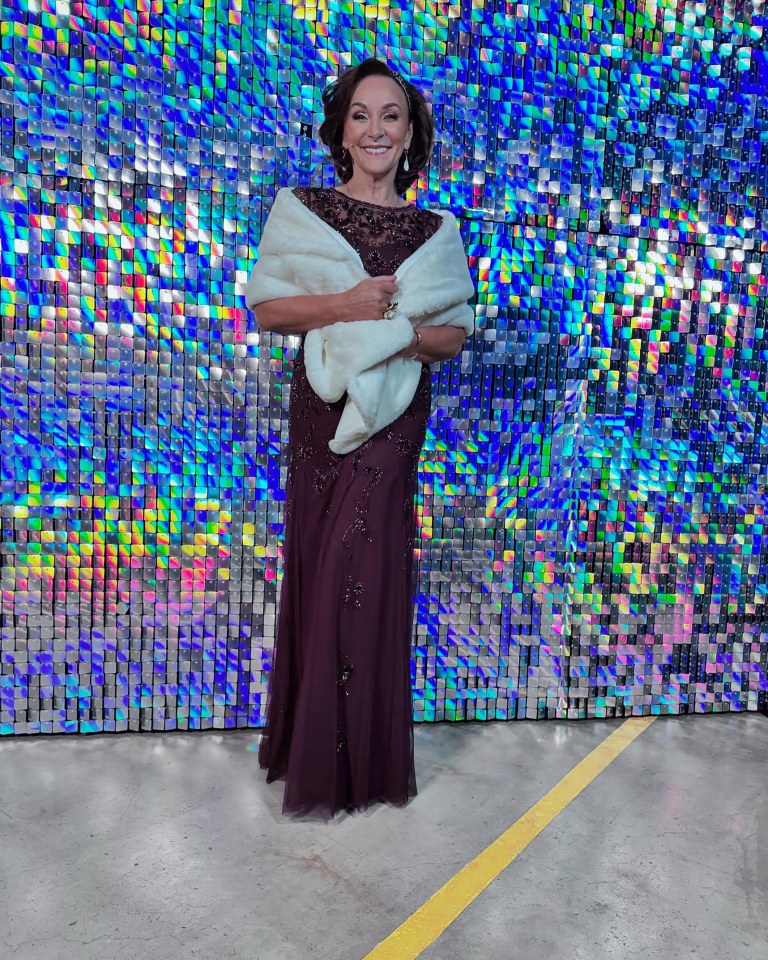 Shirley Ballas in a burgundy gown and white fur stole, posing in front of a shimmering background.