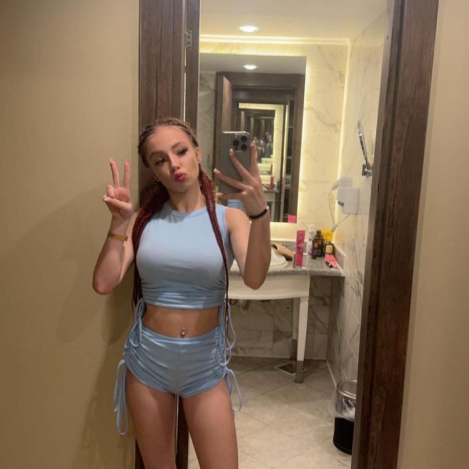 A young woman in a light blue crop top and shorts takes a selfie.