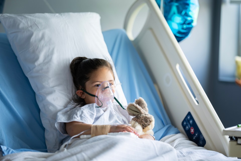 Young girl in hospital bed with oxygen mask and stuffed animal.