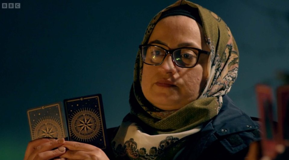 A woman wearing glasses and a headscarf holds two tarot cards.