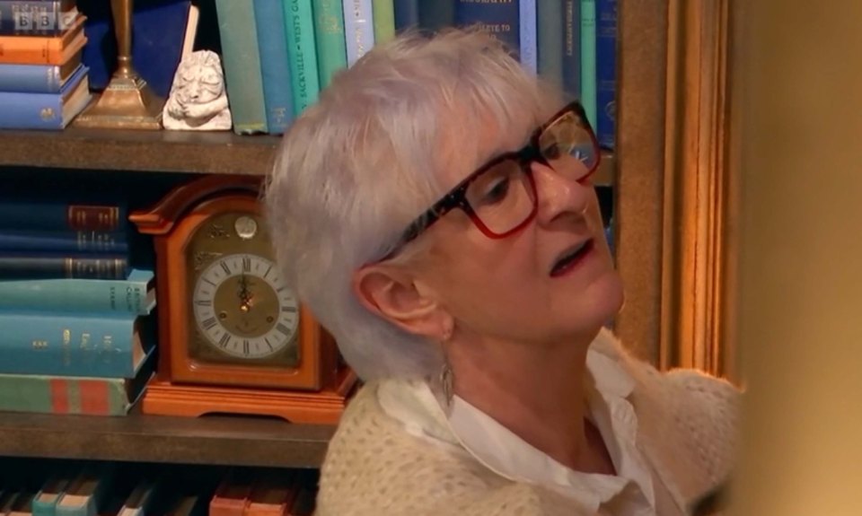 A woman with short, light-grey hair and red glasses reacts to something.