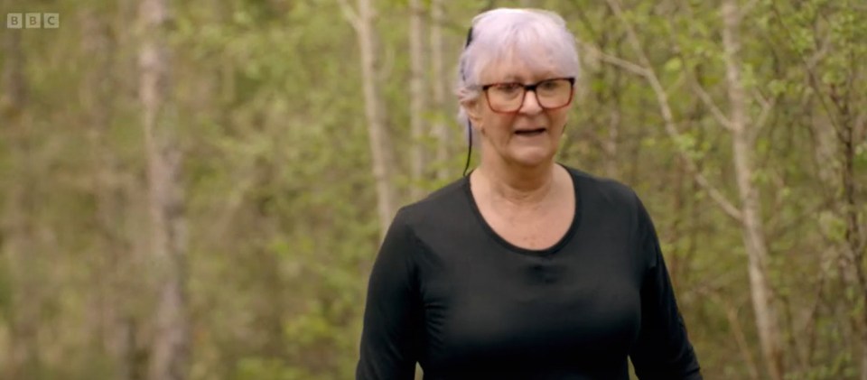 A woman in a black shirt and glasses stands in a wooded area, looking serious.  Traitors Ep2.