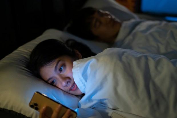 Woman using smartphone in bed while partner sleeps.