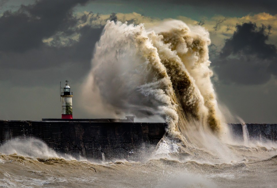 Large waves crashing over a jetty with a lighthouse during a storm.