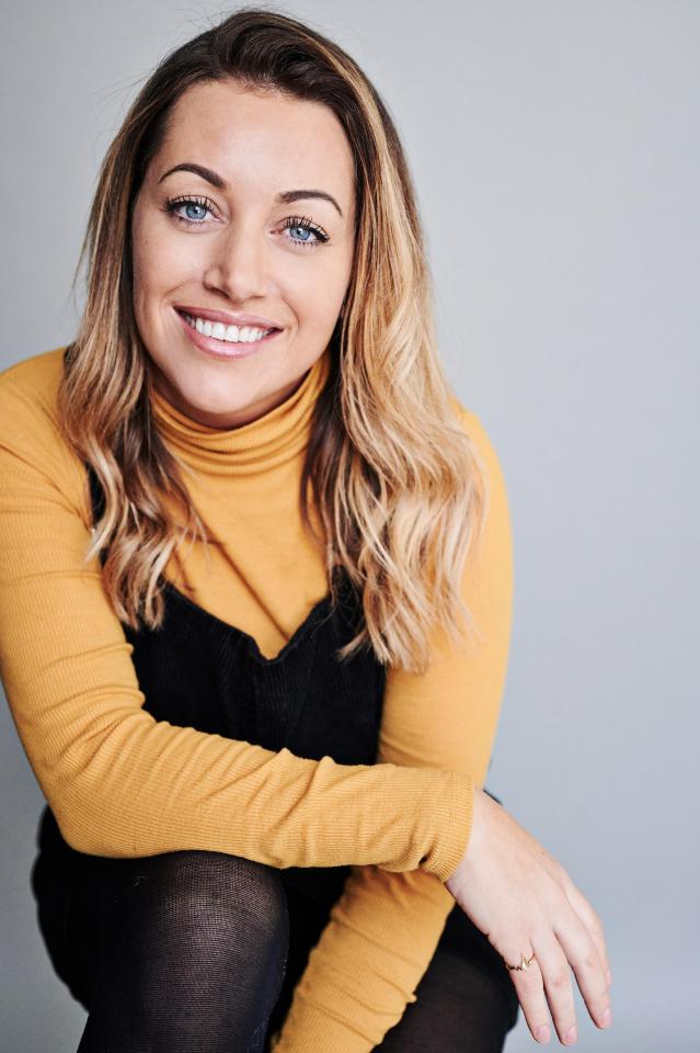 Portrait of a smiling woman with blonde hair wearing a mustard-colored turtleneck and black overalls.