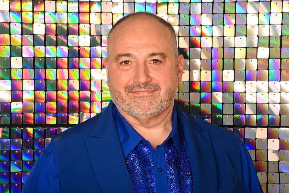 Wynne Evans in a blue blazer and sequined shirt, against a shimmering background.