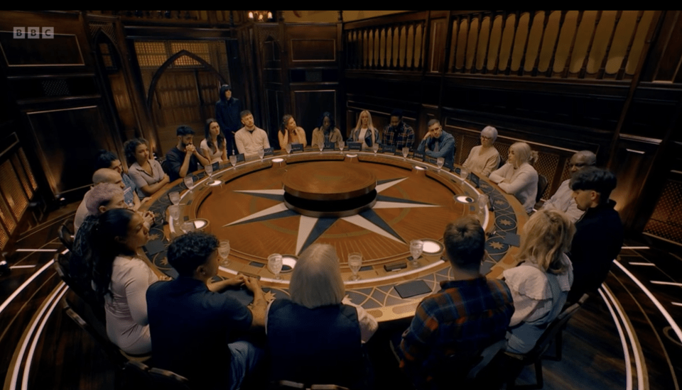 A group of people sitting around a large circular table in a room with dark wood paneling.