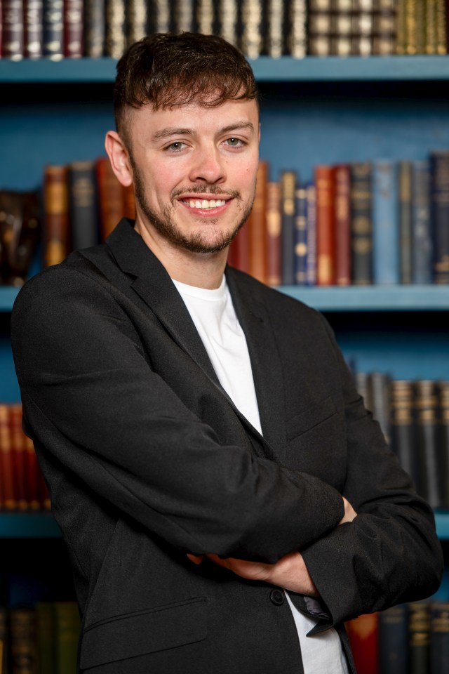 Portrait of a man in a black blazer, arms crossed, standing in front of bookshelves.