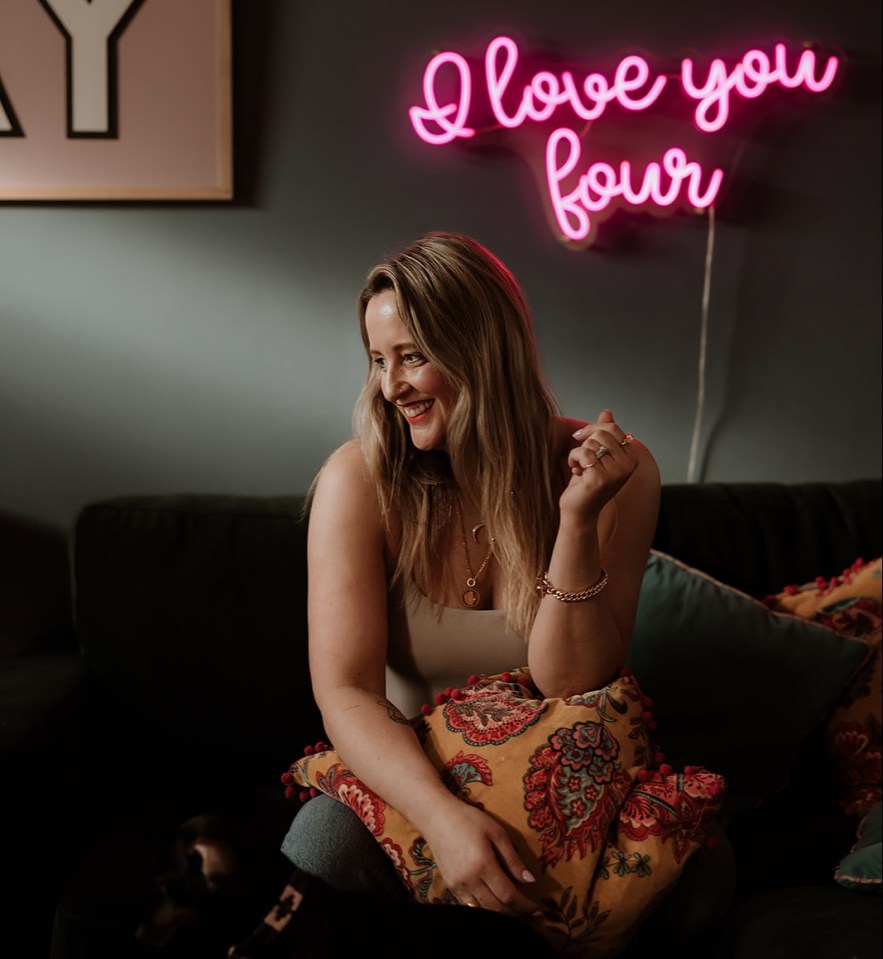 Woman sitting on a couch with a dog, smiling at something off-camera.  A pink neon sign reading "I love you four" is on the wall behind her.