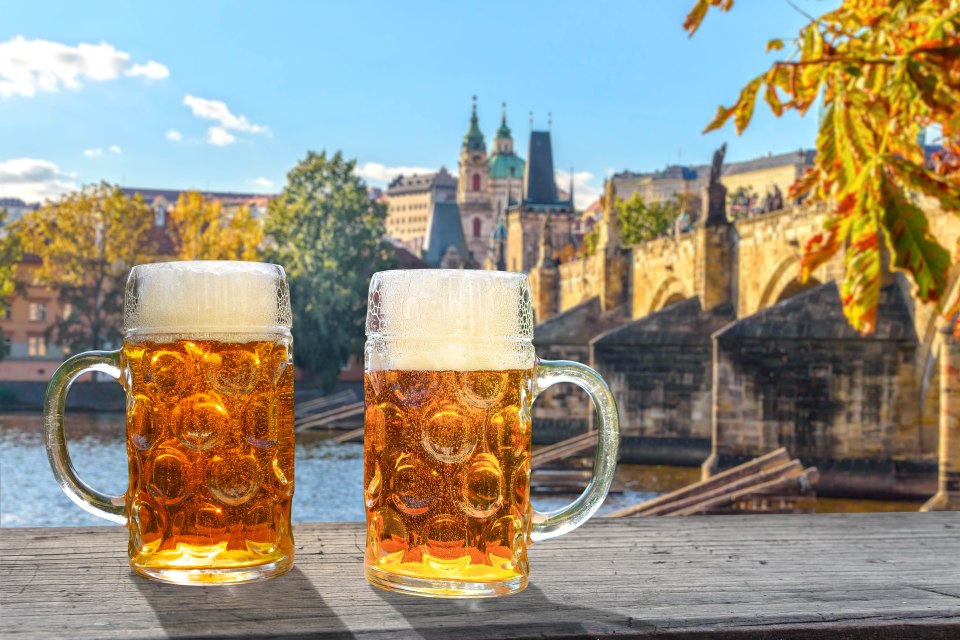 Two beer steins on a wooden surface with a view of Prague in the background.