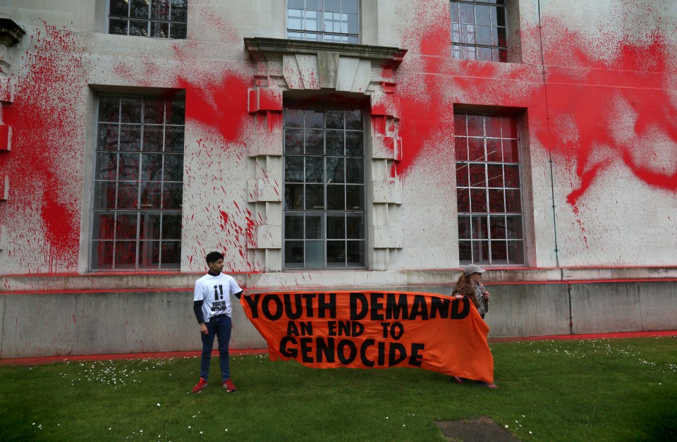 Two young people holding a banner that reads "Youth Demand an End to Genocide" in front of a building splattered with red paint.