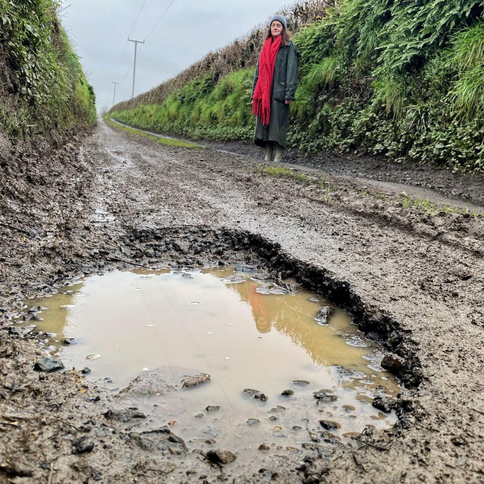 Woman standing near a large pothole in a muddy road.