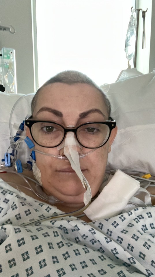 A woman with a shaved head, wearing glasses and a nasal cannula, lies in a hospital bed.