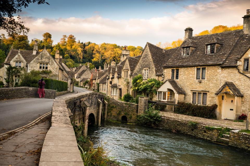 A woman and child walk across a stone bridge over a river in a quaint village.