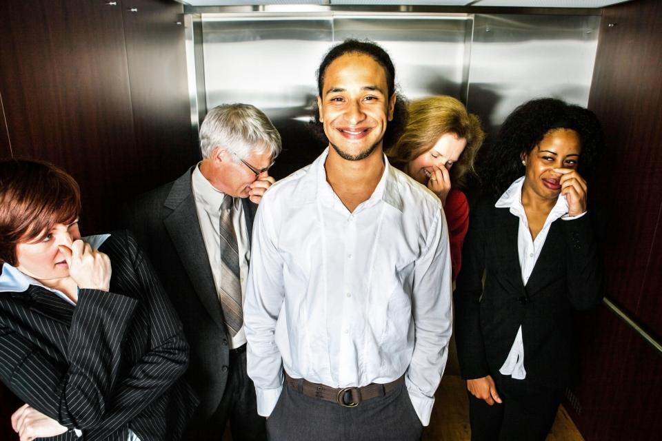 A young man smiles awkwardly in an elevator as other passengers hold their noses.