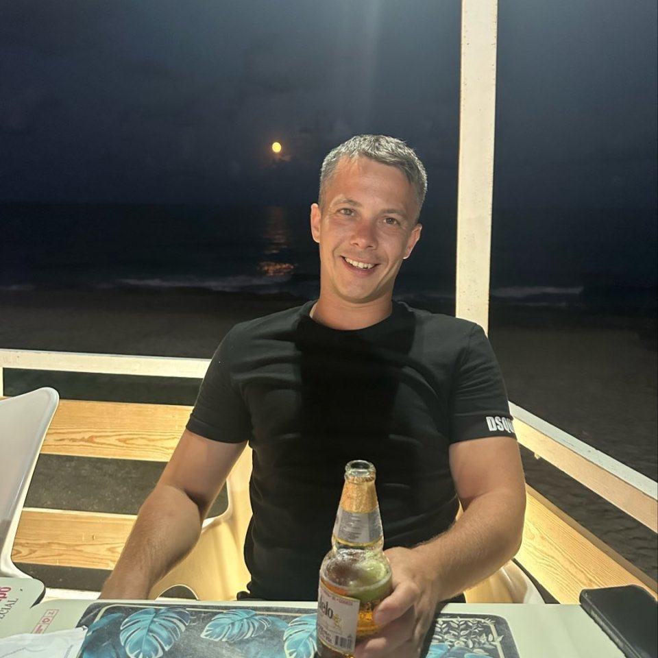 Man holding a beer bottle at an outdoor beachfront restaurant at night.