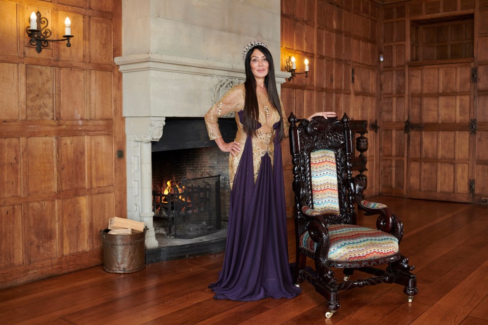 Woman in a crown and long gown standing by a fireplace and ornate chair.